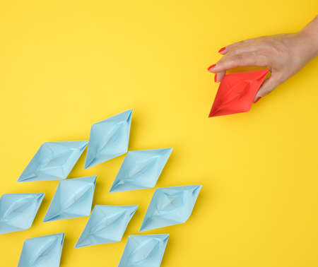 a group of blue paper boats follows a red boat in front of a yellow background. The concept of a...
