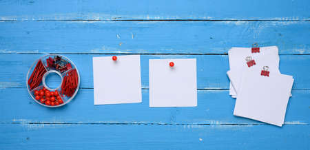 blank square paper sheets and stationery set with paper clips and clips on blue wooden background