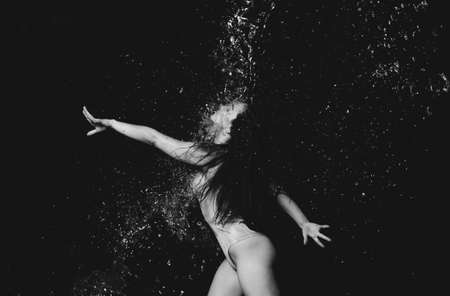 young beautiful woman of Caucasian appearance with long hair dances in drops of water on a black background. Jumping and swinging arms, dressed in a beige bodysuitの写真素材