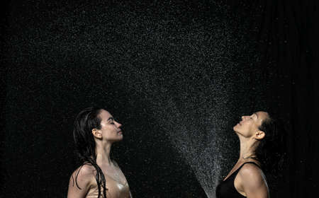 two beautiful women of Caucasian appearance with black hair sit in drops of water on a black...