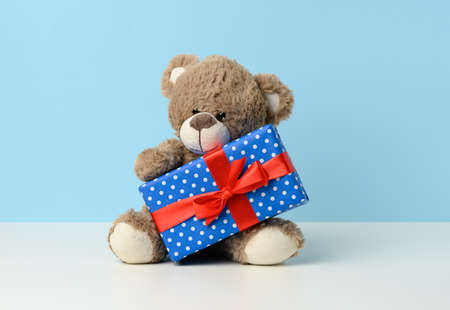 cute brown teddy bear holding a box wrapped in blue paper and red silk ribbon on white table. Prize and congratulationsの写真素材