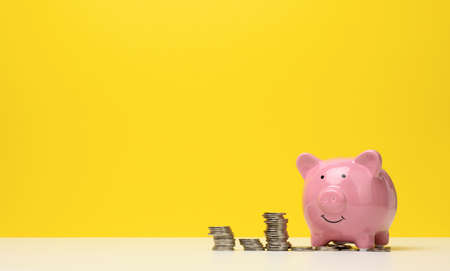 pink ceramic piggy bank and a stack of coins on a white table, yellow background. Savings concept,...
