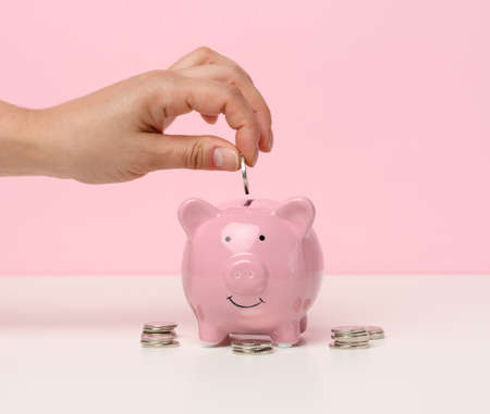female hand throws a coin into a pink piggy bank on a white table. Concept of accumulating cash,...