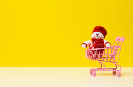 textile snowman in a metal miniature shopping cart on a white table, yellow background. Festive background for Christmas and New Year, copy spaceの写真素材