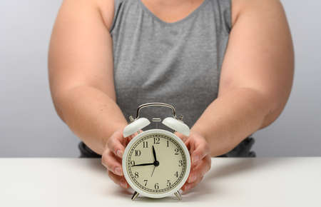 plump woman sits at a table and holds a round white alarm clock. The concept of fractional nutrition, time to lose weight, adherence to sleepの写真素材