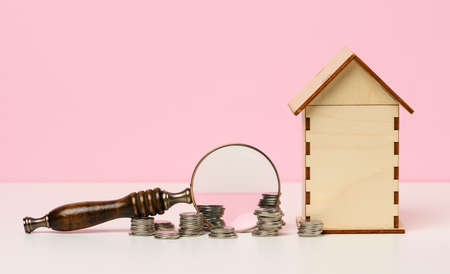 stack of metallic money and magnifier and wooden house on a pink background. Real estate rental, purchase and sale concept. Realtor services, building repair and maintenanceの写真素材