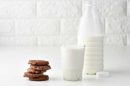 a full glass cup of milk and a clear plastic bottle with milk, next to a stack of round chocolate chip cookies on a white tableの写真素材
