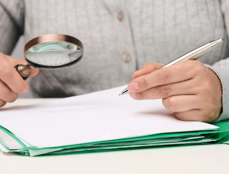 a woman sits at a white table and holds a metal pen over a pile of papers, in the other hand a...