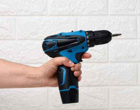 hand in a textile glove holds a portable drill on a battery against a background of a white brick wallの写真素材