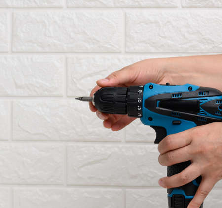 hand holds a portable drill on a battery against a background of a white brick wallの写真素材