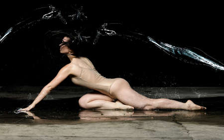 young woman with black long hair lies on the ground under raindrops on a black background. Woman dressed in beige bodysuitの写真素材