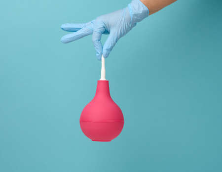 a hand in a blue medical glove holds a pink rubber enema on a blue backgroundの写真素材