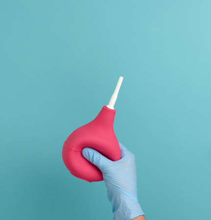 a hand in a blue medical glove holds a pink rubber enema on a blue backgroundの写真素材