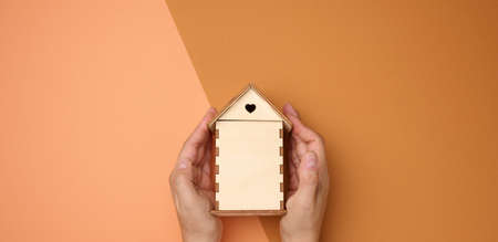 female hands folded to each other over a wooden miniature model house on a brown background. Real estate insurance concept, environmental protection, family happinessの写真素材