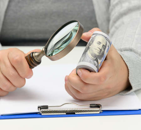 a stack of one hundred dollar bills in a woman's hand and a wooden magnifying glass on a white...