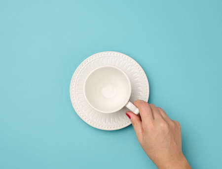 female hand holding a white ceramic mug with a saucer on a blue background, top view