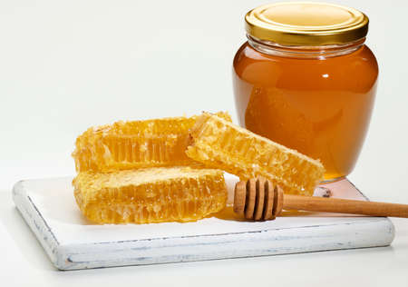 glass transparent jar with honey and pieces of wax honeycomb with honey on a white wooden board,...