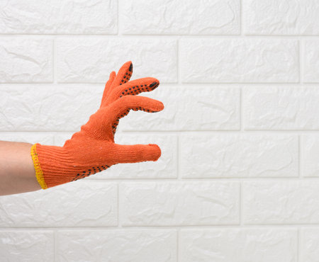 female hand in an orange work protective glove against the background of a white brick wall, the hand conditionally holds an objectの写真素材