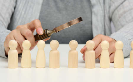 figurines of men on a white table, a female hand holds a magnifying glass over one. concept of searching for employees in the company, recruiting personnel, identifying talented and strong personalitiの写真素材