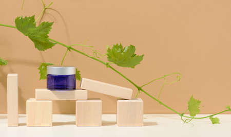 cosmetic products in a blue glass jar with a gray lid on a wooden podium made of cubes, behind a branch of grapes with green leaves. Blank for branding products, moisturizer on beige backgroundの写真素材