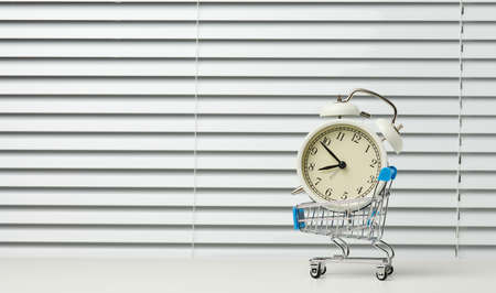 miniature metal shopping cart on wheels and in the middle of a round alarm clock on a white table. Start of discounts, sale, copy spaceの写真素材
