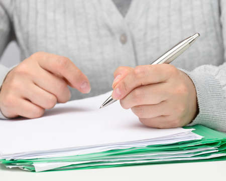 woman of Caucasian appearance sits in gray clothes at a white table and signs documents with a metal...
