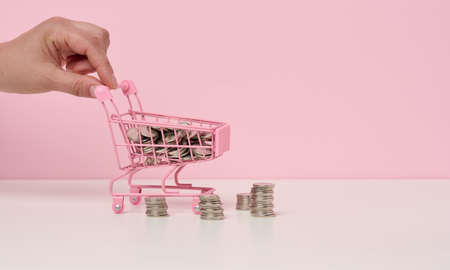female hand pushes a miniature metal shopping cart and a stack of coins on a white table. The concept of discounts and sales, budget savings. Online tradeの写真素材