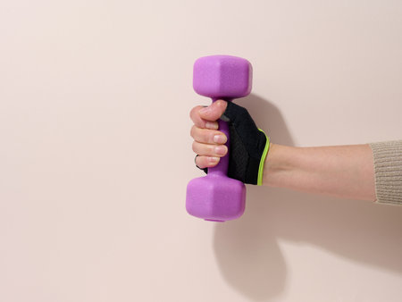 female hand in a black sports glove holds a purple one kilogram dumbbell on a beige backgroundの写真素材