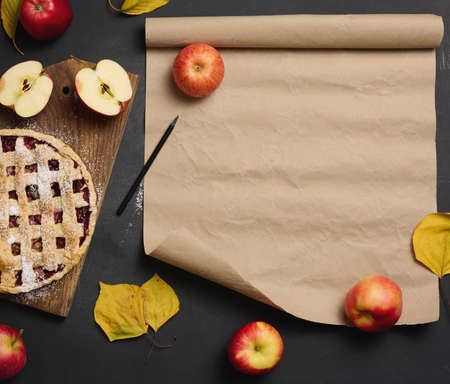 baked round traditional apple pie on a brown wooden board and fresh red apples. Nearby is a roll of brown paper for writing a recipe or menu, top viewの写真素材