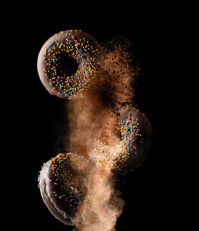 chocolate round donuts with multicolored sugar sprinkles levitate in a cloud of brown cocoa on a black background. Powder flies upの写真素材