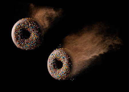 chocolate round donuts with multicolored sugar sprinkles levitate in a cloud of brown cocoa on a black backgroundの写真素材