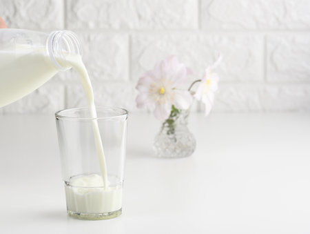 process of pouring fresh milk from a plastic bottle into a glass cup, white tableの写真素材
