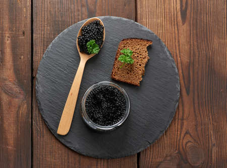 black paddlefish caviar in a wooden spoon, a slice of bread and a jar on a black round graphite board, top viewの写真素材
