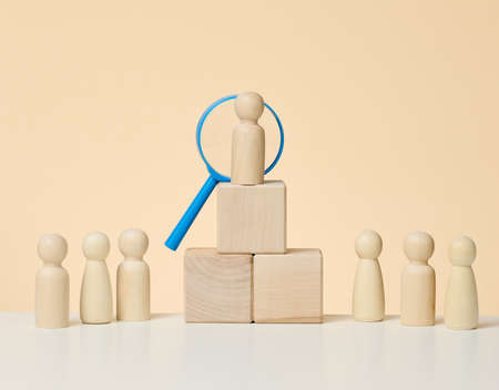 wooden figures of men stand on a beige background and a blue plastic magnifying glass. Recruitment concept, search for talented and capable employees, career growthの写真素材