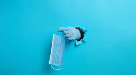 a female hand holds a disposable medical mask to protect against viruses during an epidemic and pandemic. Part of the body sticks out of a torn hole in blue paperの写真素材