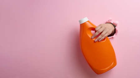 orange plastic bottle with liquid detergent in a female hand on a pink background. A part of the body sticks out of a torn hole in the background, a place for an inscriptionの写真素材