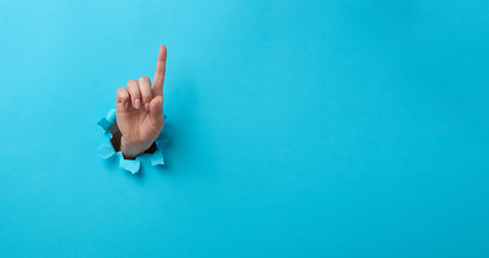 female hand sticking out of a torn hole in a blue paper background, attention gesture. Place for an inscription, bannerの写真素材
