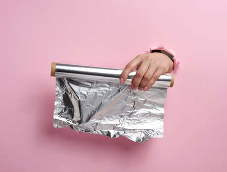 female hand holding a scroll of gray food foil on a pink background, part of the body sticking out of a torn holeの写真素材