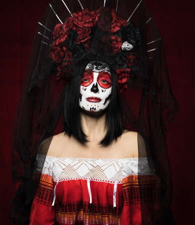 portrait of a beautiful woman with a sugar skull makeup with a wreath of flowers on her head, red backgroundの写真素材