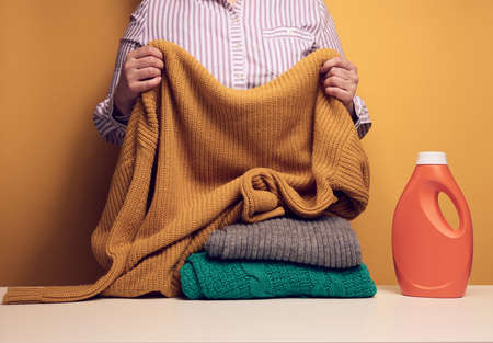 adult woman folds washed dry clothes on a white table, next to a plastic bottle with liquid washing gel, yellow backgroundの写真素材