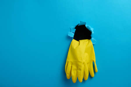 a pair of yellow latex house cleaning gloves sticking out of the torn hole of the blue paper background, copy spaceの写真素材