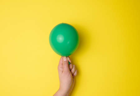 female hand holding an inflated green air balloon on a yellow background, close upの写真素材