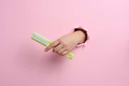 female hand holds a green plastic brush on a pink background. A part of the body sticks out of a hole with torn edges in a paper backgroundの写真素材