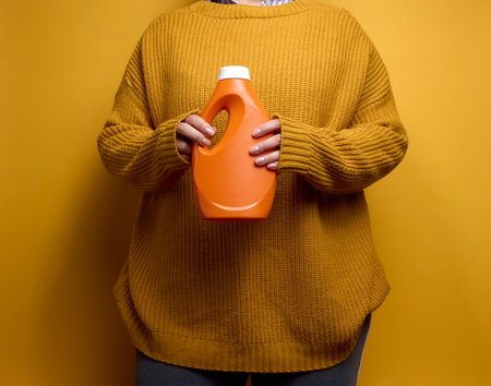 woman in a knitted sweater holds an orange plastic bottle with liquid washing gel. Laundry and chores, yellow backgroundの写真素材