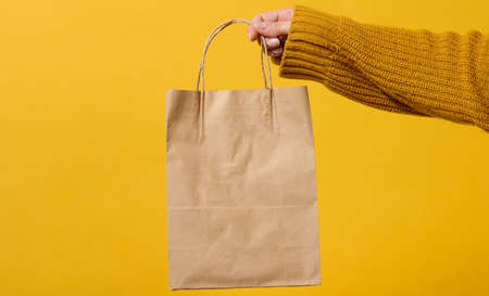 female hand in a sweater holds a brown craft paper bag with handles on a yellow background. Zero wasteの写真素材