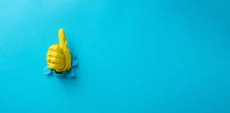 a hand in a yellow household rubber glove sticks out of a torn hole and shows a gesture okay, thumb...