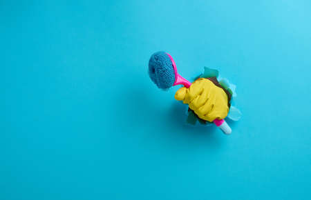 hand in a yellow rubber glove holds a plastic cleaning brush on a blue background, copy space
