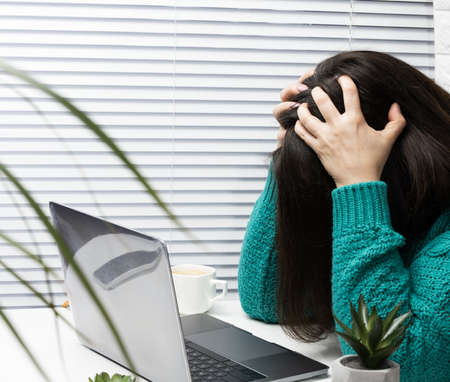 a woman in a sweater sits at the table with her head clasped in her hands, stress and headache from...