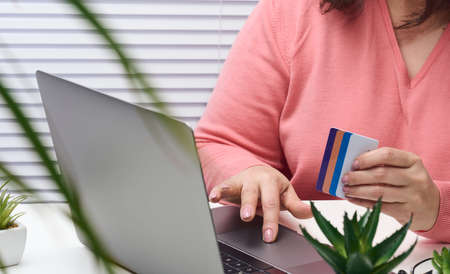 woman in a pink sweater makes purchases online with a laptop, a credit card in hand. Woman sitting at a white table and looking at the monitorの写真素材