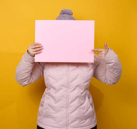 woman in pink winter jacket and hat holds blank pink sheet of paper on yellow background. Seasonal sale. Place for inscriptionの写真素材